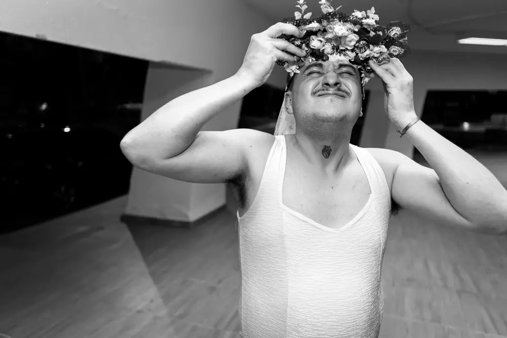 En blanco y negro, un participante se acomoda una corona de flores con vestido de novia
