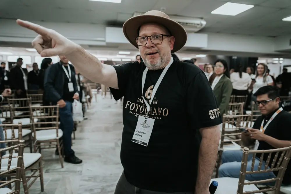 Un fotógrafo con sombrero señala a alguien del público en el pasillo del salón